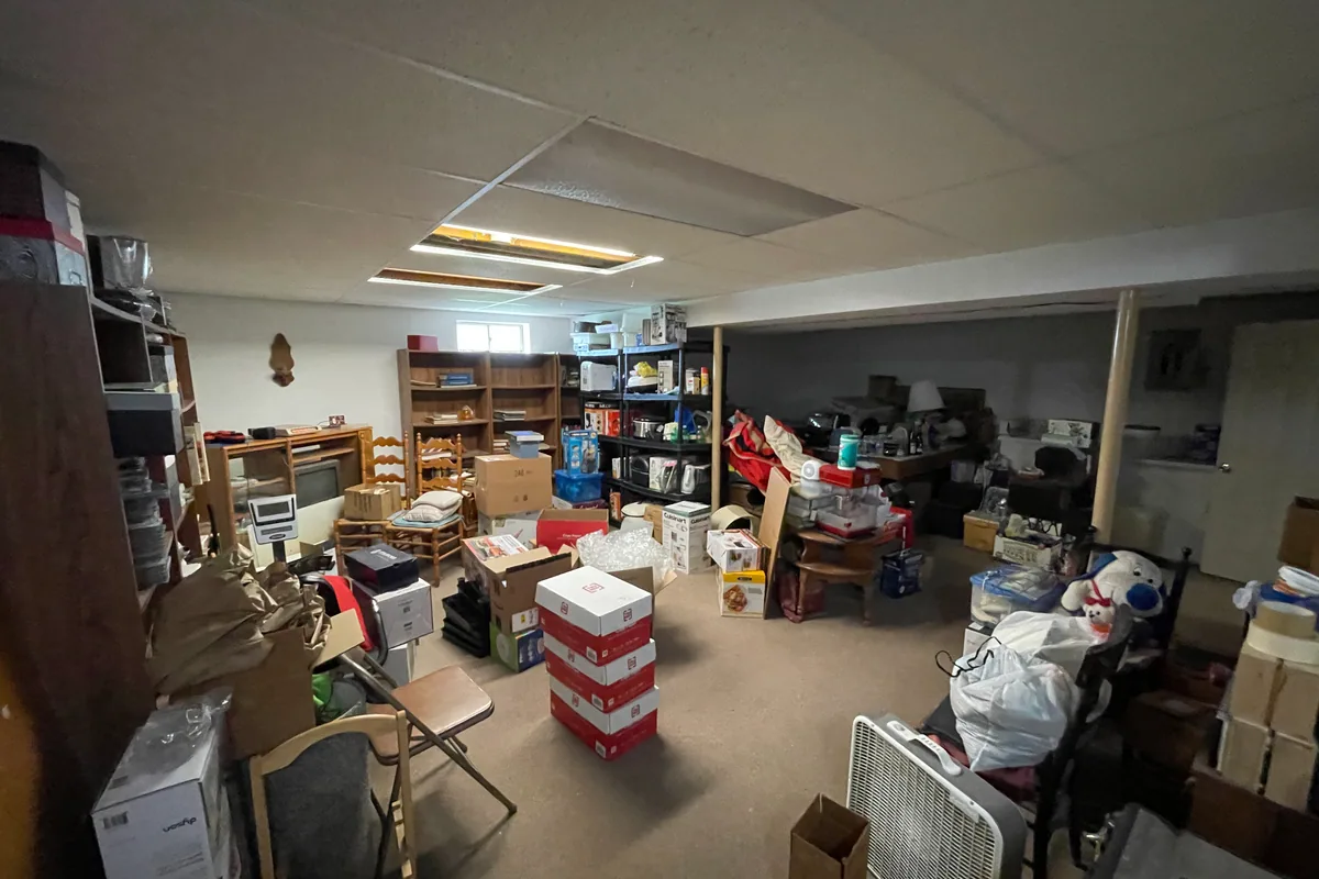 Huber Heights Chukar Drive basement before sale — cluttered storage with bins, folding chairs, sagging ceiling tiles, matted carpet