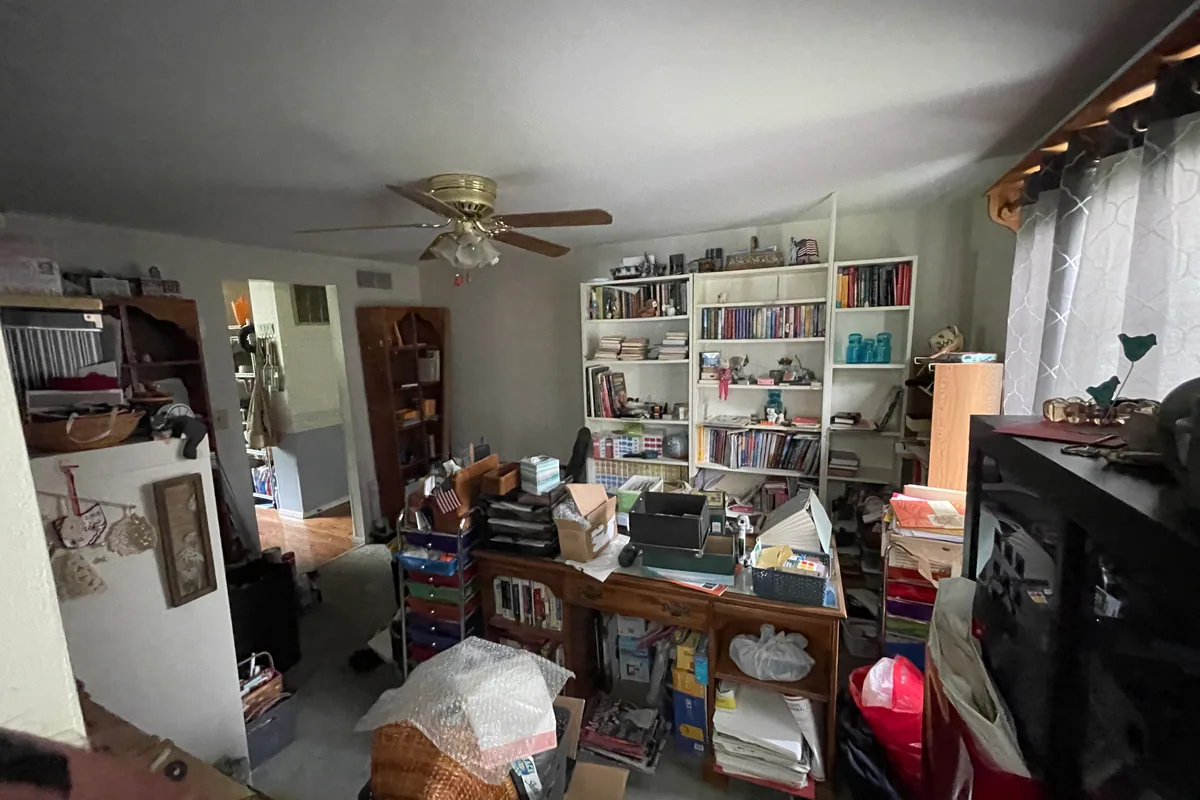Huber Heights Chukar Drive room before sale — built-in white bookshelves stuffed with books, cluttered roll-top desk, brass ceiling fan