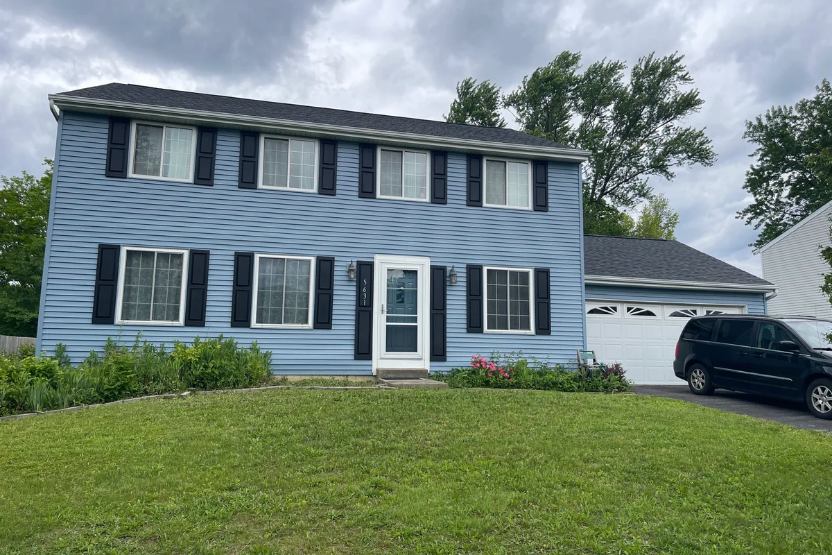 Huber Heights Chukar Drive two-story colonial before cash sale to EZ Sell Homebuyers — faded blue siding, weedy beds, dated front door