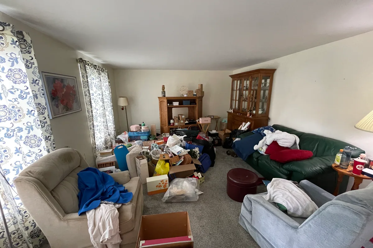Huber Heights Chukar Drive living room before sale — cluttered with belongings, dated furniture, worn carpet, blue patterned curtains