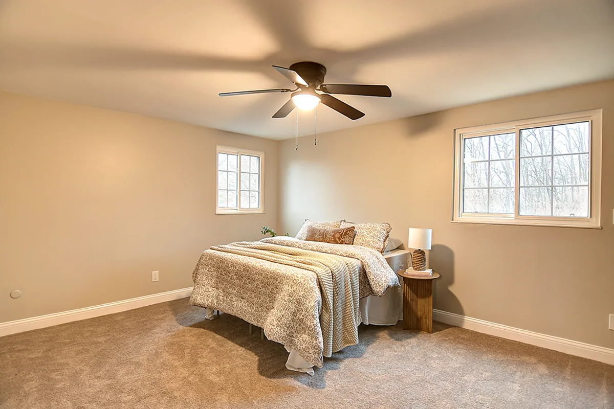 Renovated Huber Heights master bedroom after EZ Sell Homebuyers — warm neutral paint, fresh carpet, updated bronze ceiling fan, staged with bed and nightstands, bright natural light from two window walls