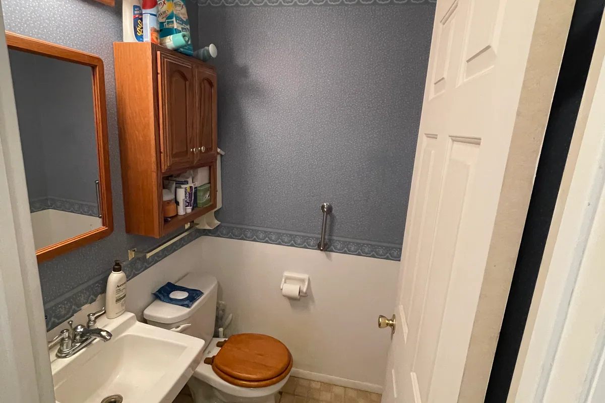 Huber Heights Chukar Drive powder room before sale — blue floral wallpaper, oak wall cabinet, dated pedestal sink, wooden toilet seat, vinyl tile floor
